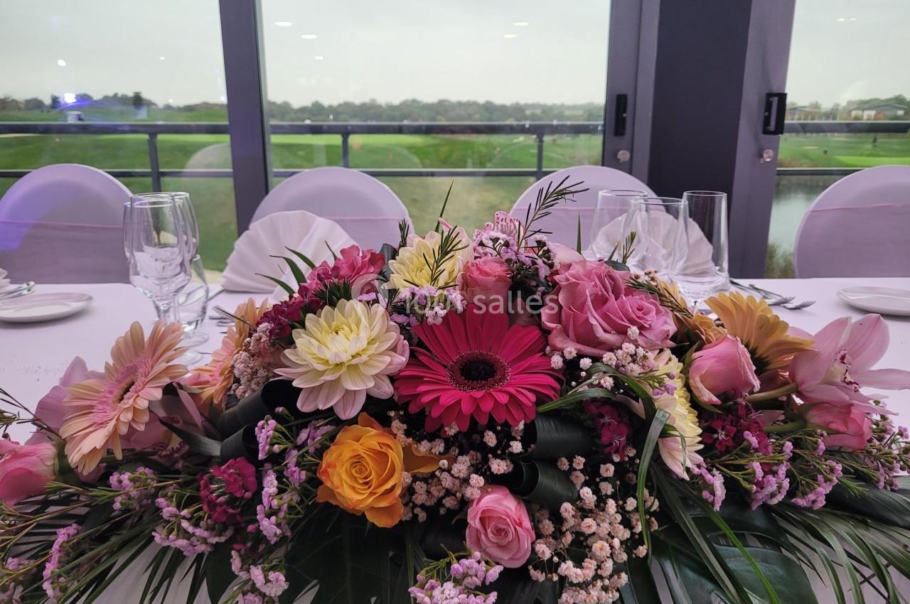 Centre de table floral coloré avec des roses, gerberas et feuillage, disposé sur une table blanche près d'une baie vitrée.