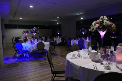 Salle de réception décorée avec des tables dressées, des chaises ornées de nœuds roses et des bouquets de fleurs.