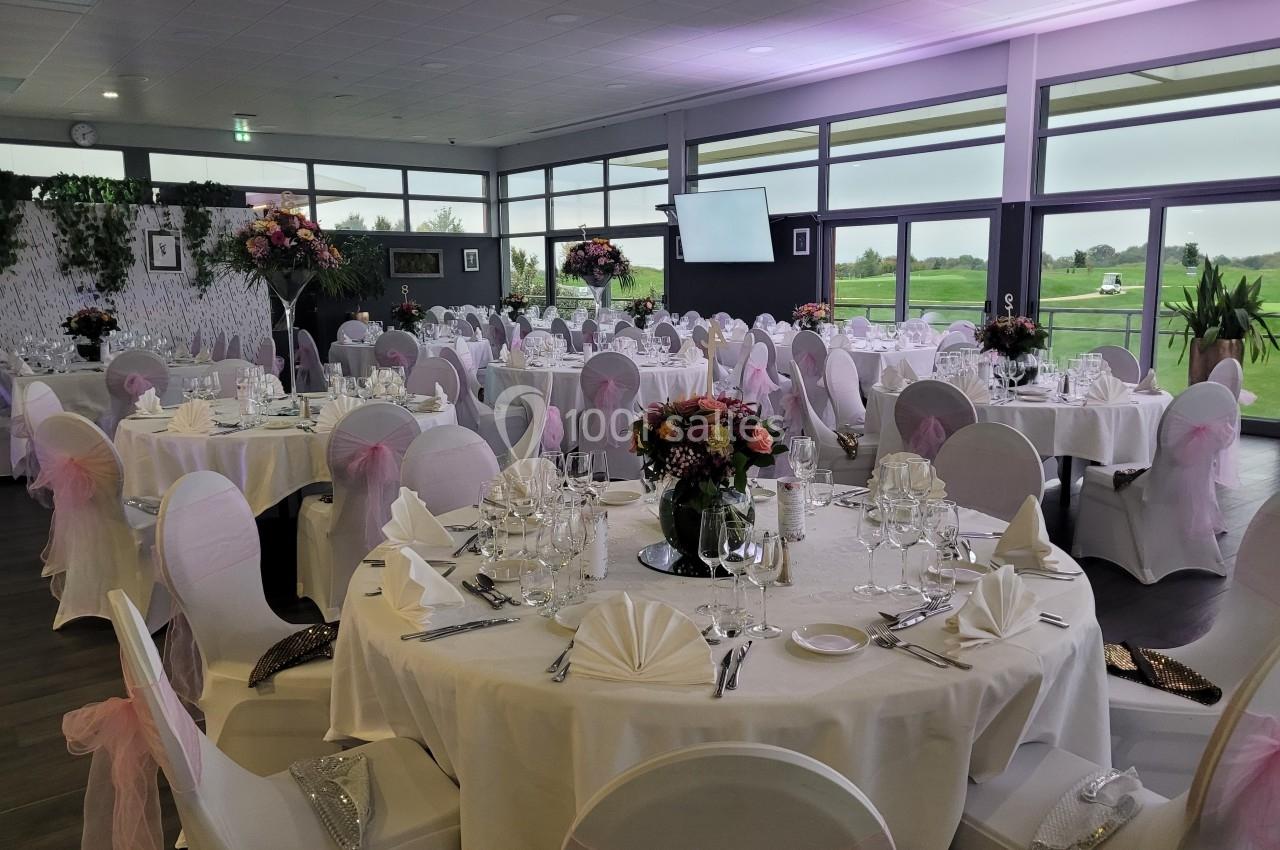 Salle de réception décorée avec des tables rondes, nappes blanches, chaises ornées de nœuds roses et arrangements floraux.