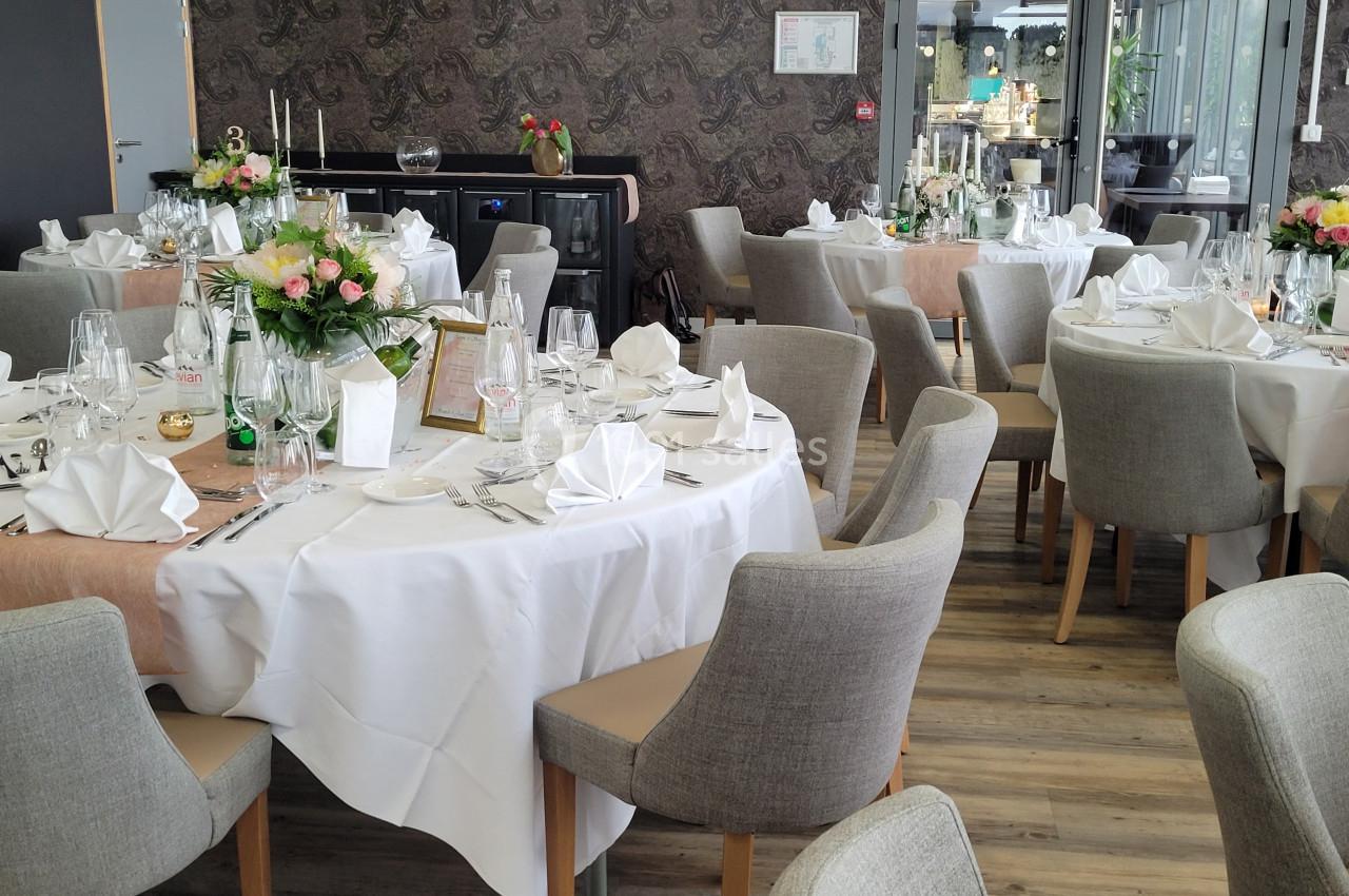 Salle de réception avec tables rondes dressées, nappes blanches, serviettes pliées et décorations florales.