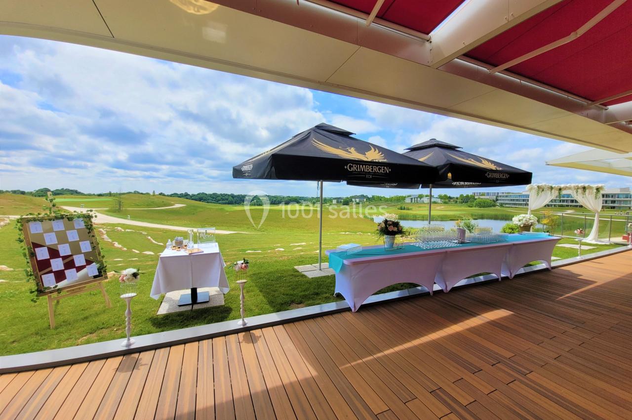 Terrasse avec tables dressées, parasols noirs et vue sur un paysage verdoyant sous un ciel partiellement nuageux.