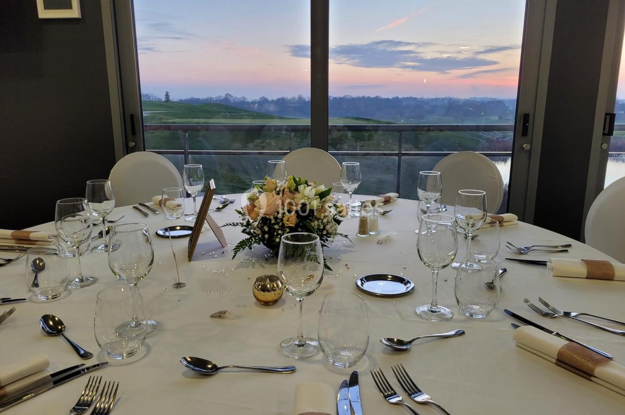 Table élégamment dressée avec des couverts, verres et centre de table floral, devant une baie vitrée au coucher du soleil.