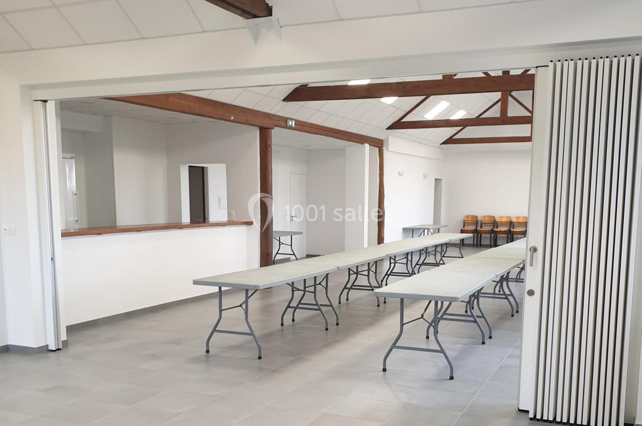 Salle lumineuse avec tables pliantes alignées, chaises empilées au fond et poutres apparentes au plafond.