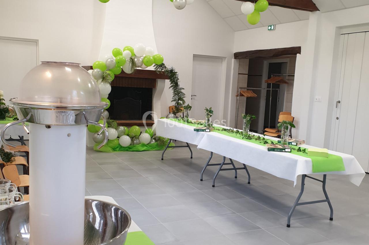 Salle décorée pour une réception, avec tables dressées, nappes vertes et blanches, et ballons autour d'une cheminée.
