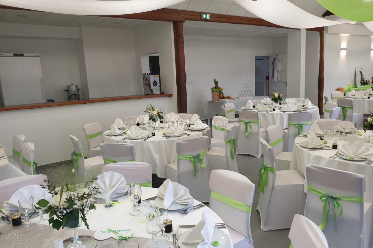 Salle de réception décorée avec des tables rondes dressées, nappes blanches et rubans verts sur les chaises.