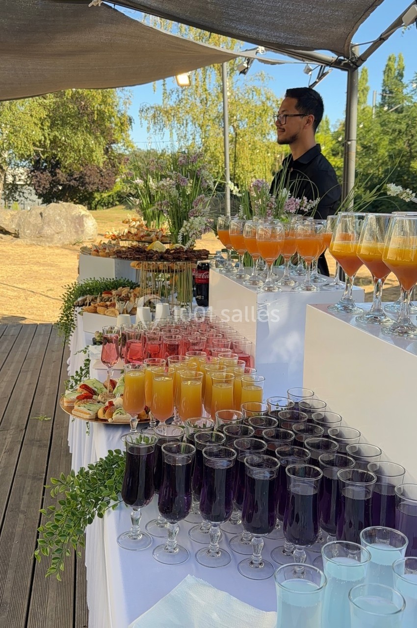 Table dressée en extérieur avec une variété de boissons colorées et d'amuse-bouches, un serveur en arrière-plan.