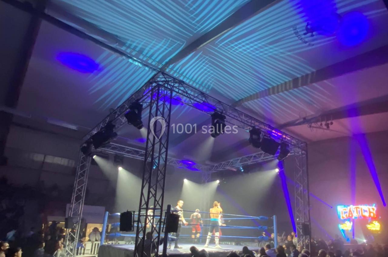 Un ring de catch éclairé par des projecteurs, entouré de spectateurs dans une salle sombre.