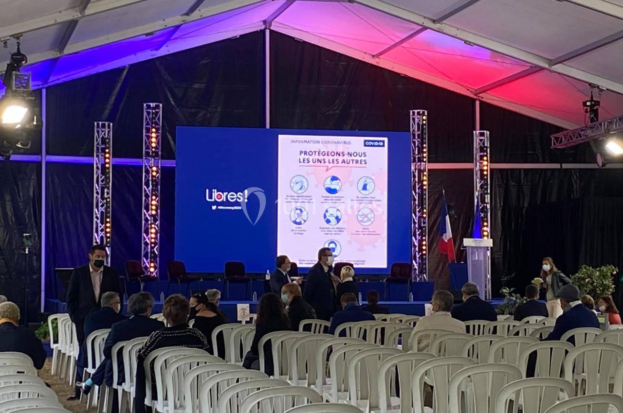 Salle de conférence sous chapiteau avec des chaises blanches, écran d'information et participants assis ou debout.