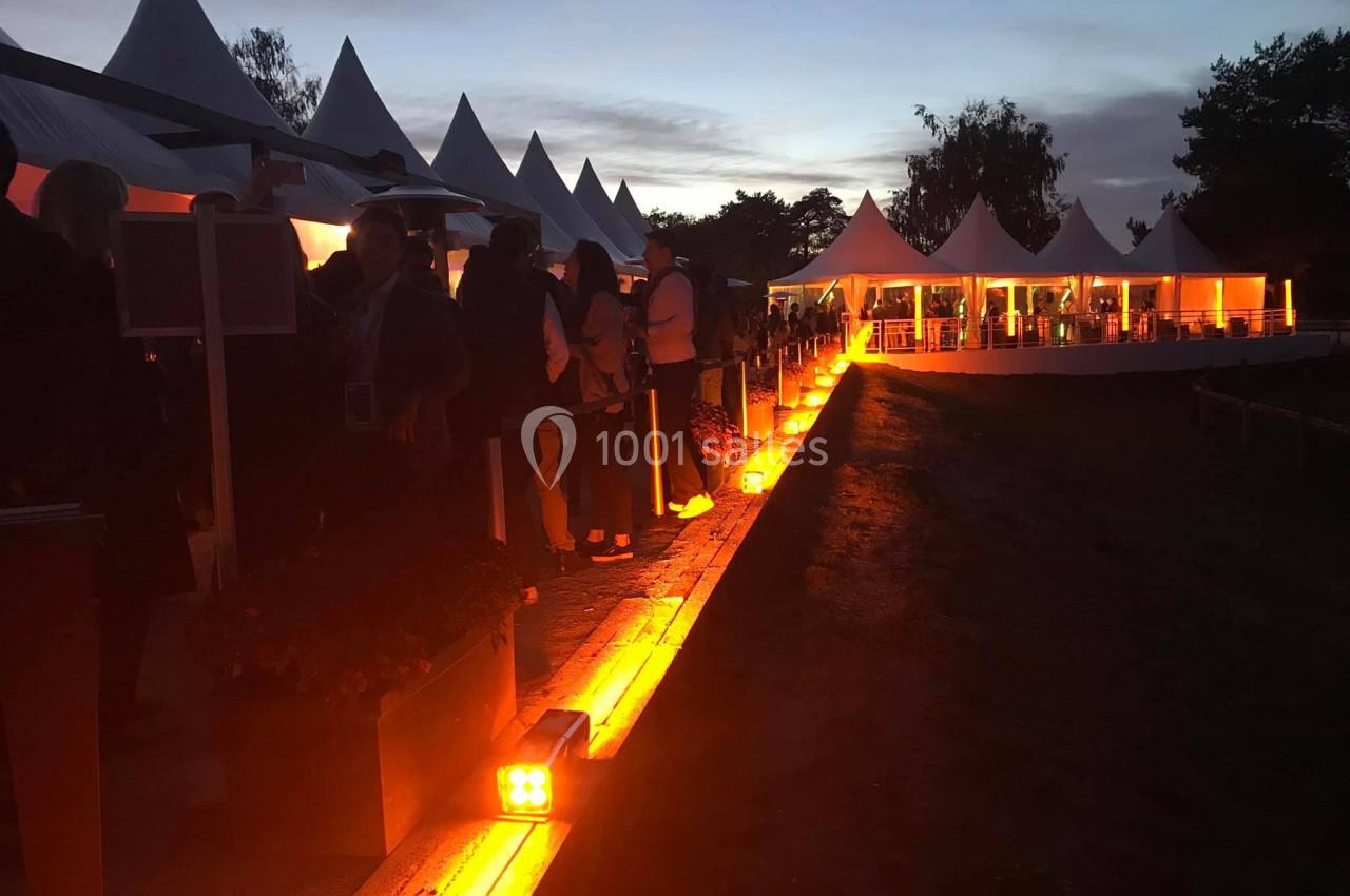 Des personnes rassemblées près de tentes blanches éclairées par des lumières orange au crépuscule.