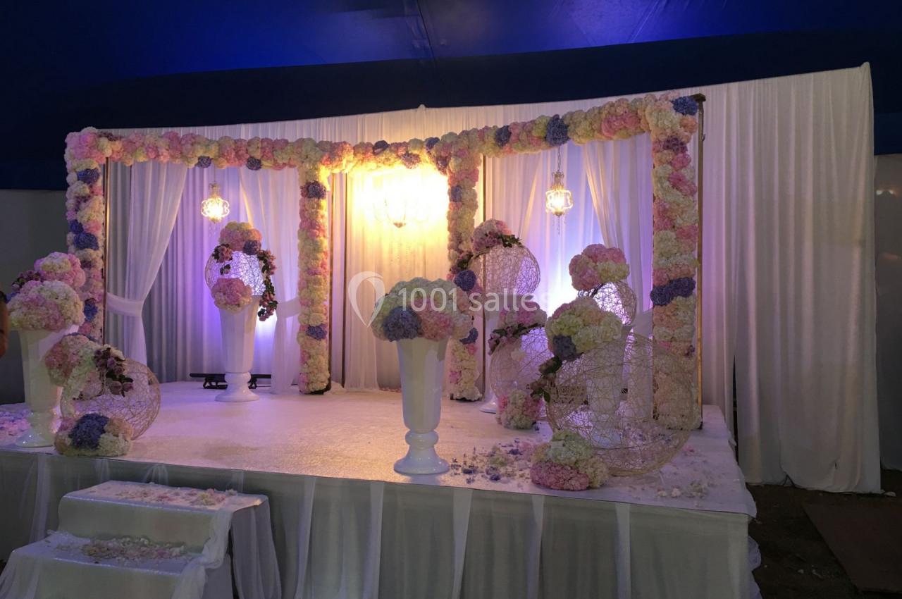 Décor de scène de mariage avec arche et colonnes ornées de fleurs pastel, éclairage doux sous une tente.