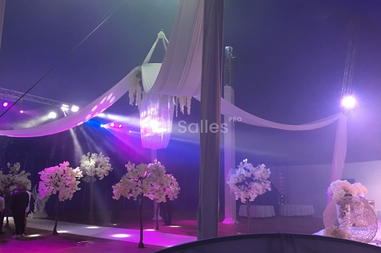 Décoration de mariage avec un lustre central, drapés blancs suspendus et arbres artificiels ornés de fleurs.