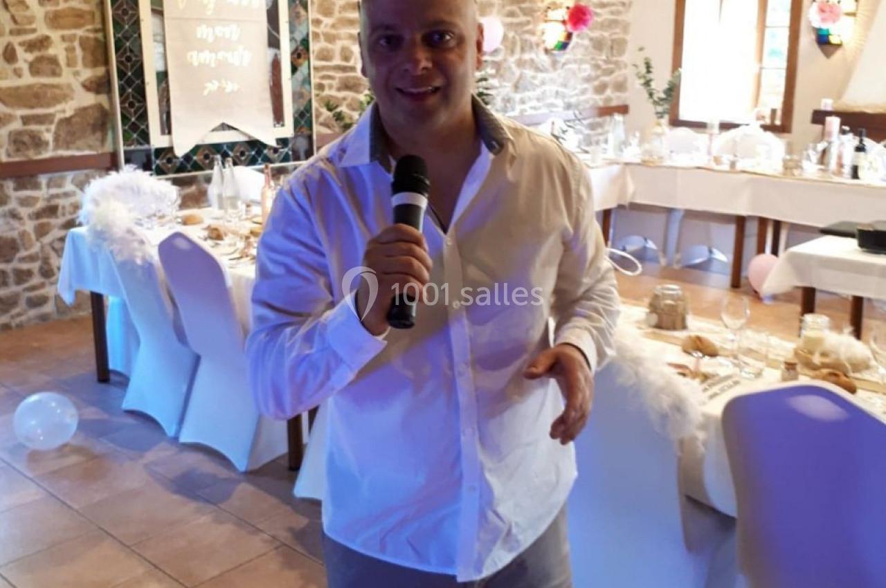 Homme debout tenant un micro dans une salle décorée pour un événement avec des tables dressées et des décorations suspendues.