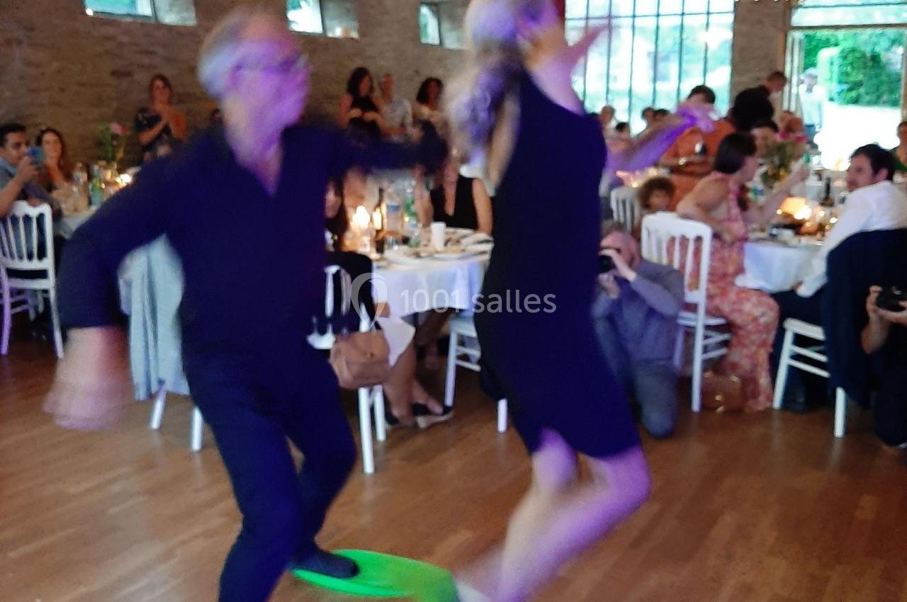 Un couple danse avec des palmes vertes dans une salle décorée de guirlandes lumineuses, devant des invités assis.