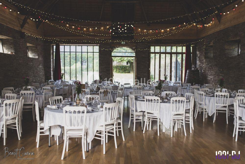 Salle de réception décorée avec des tables rondes dressées, des chaises blanches et des guirlandes lumineuses suspendues.