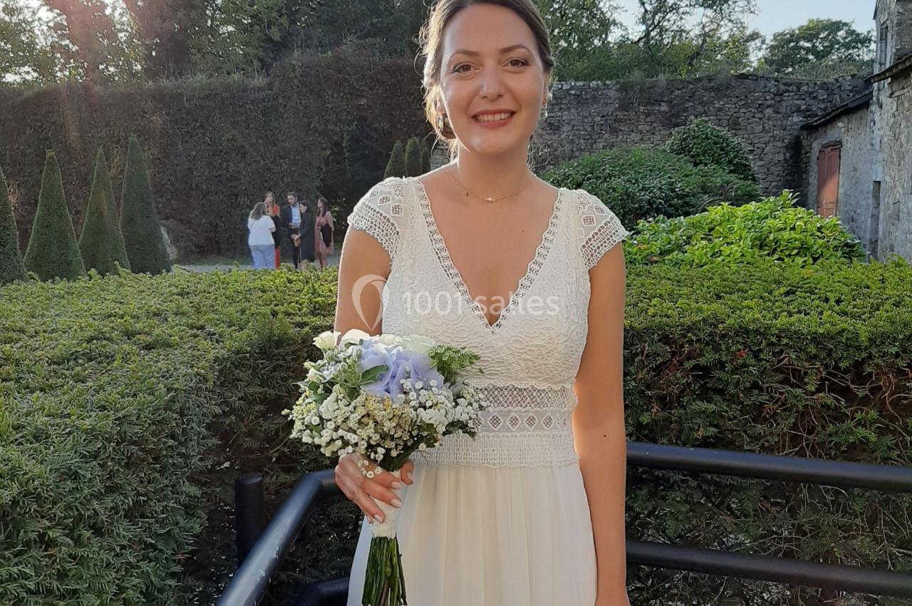 Femme en robe blanche tenant un bouquet de fleurs, debout dans un jardin avec des arbres et un mur en pierre en arrière-plan.