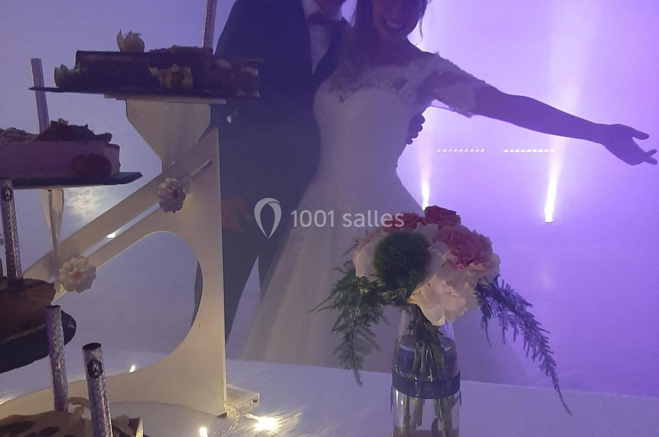 Un couple en tenue de mariage pose joyeusement devant une table décorée avec des gâteaux et un vase de fleurs.