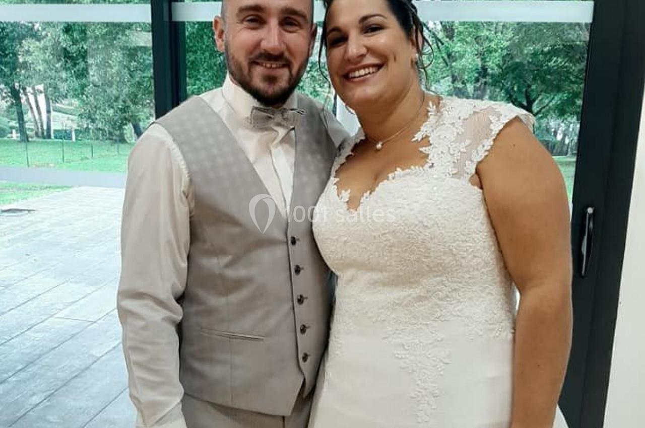 Un couple souriant en tenue de mariage pose devant une baie vitrée avec un paysage verdoyant en arrière-plan.