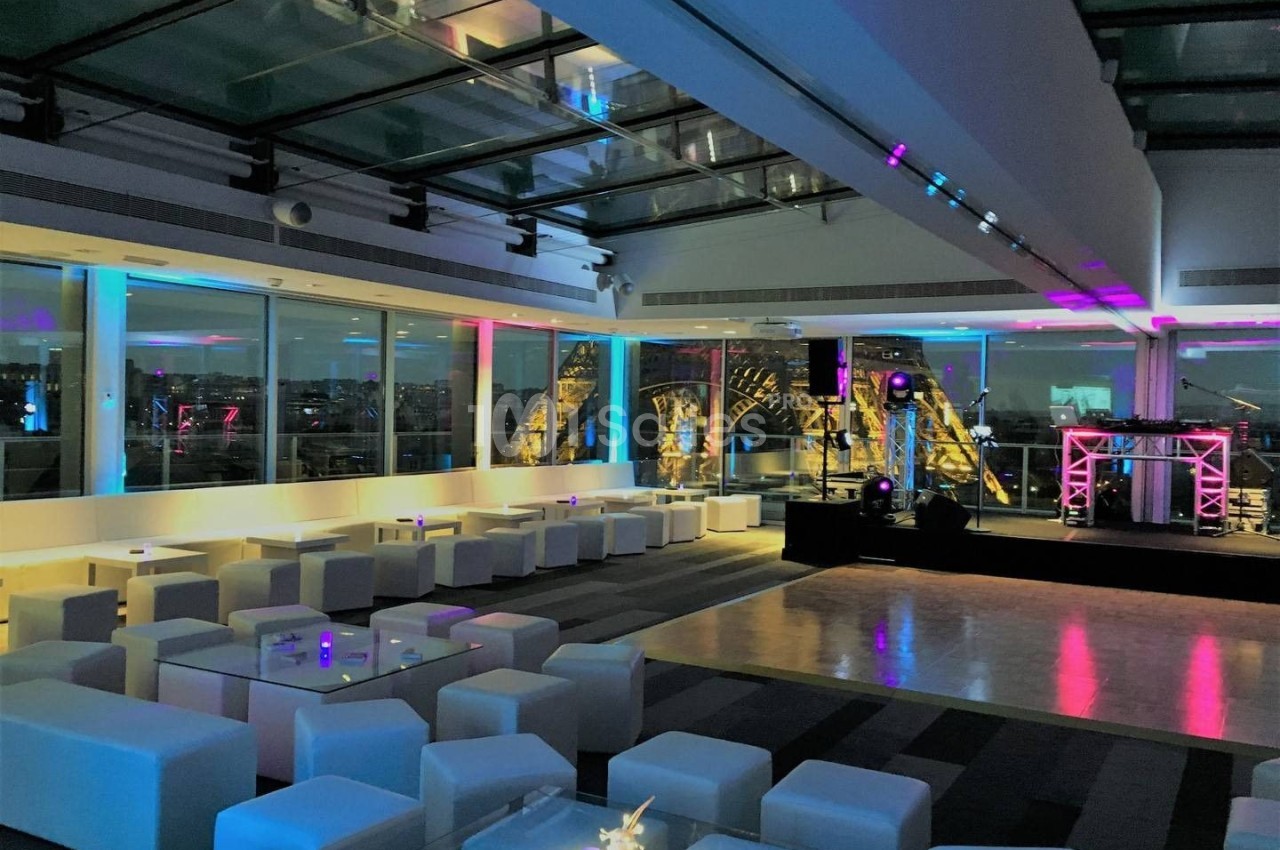 Salle moderne avec mobilier blanc, éclairage coloré et vue sur la tour Eiffel à travers de grandes baies vitrées.