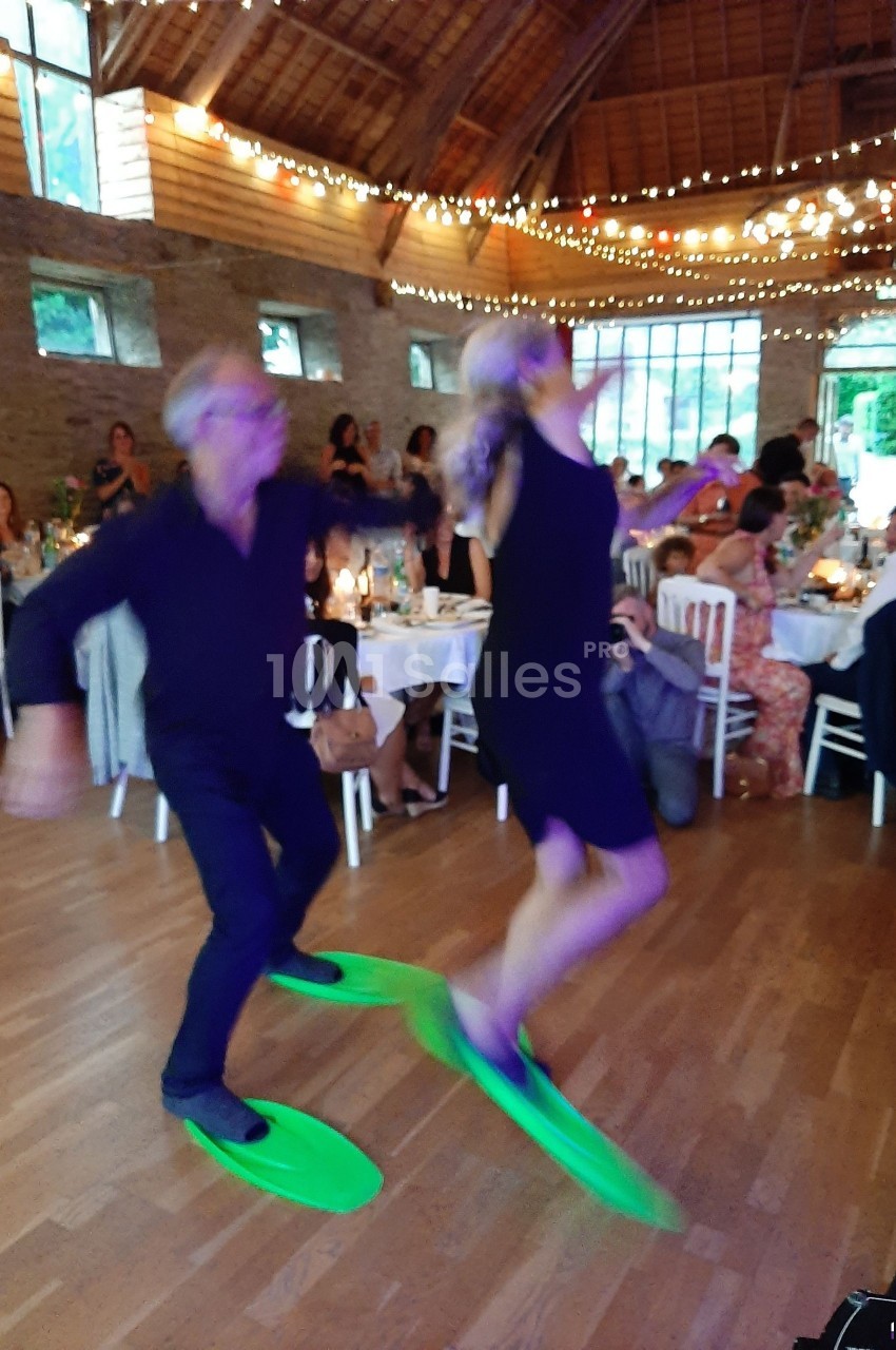 Un couple danse avec des palmes vertes dans une salle décorée de guirlandes lumineuses, devant des invités assis.