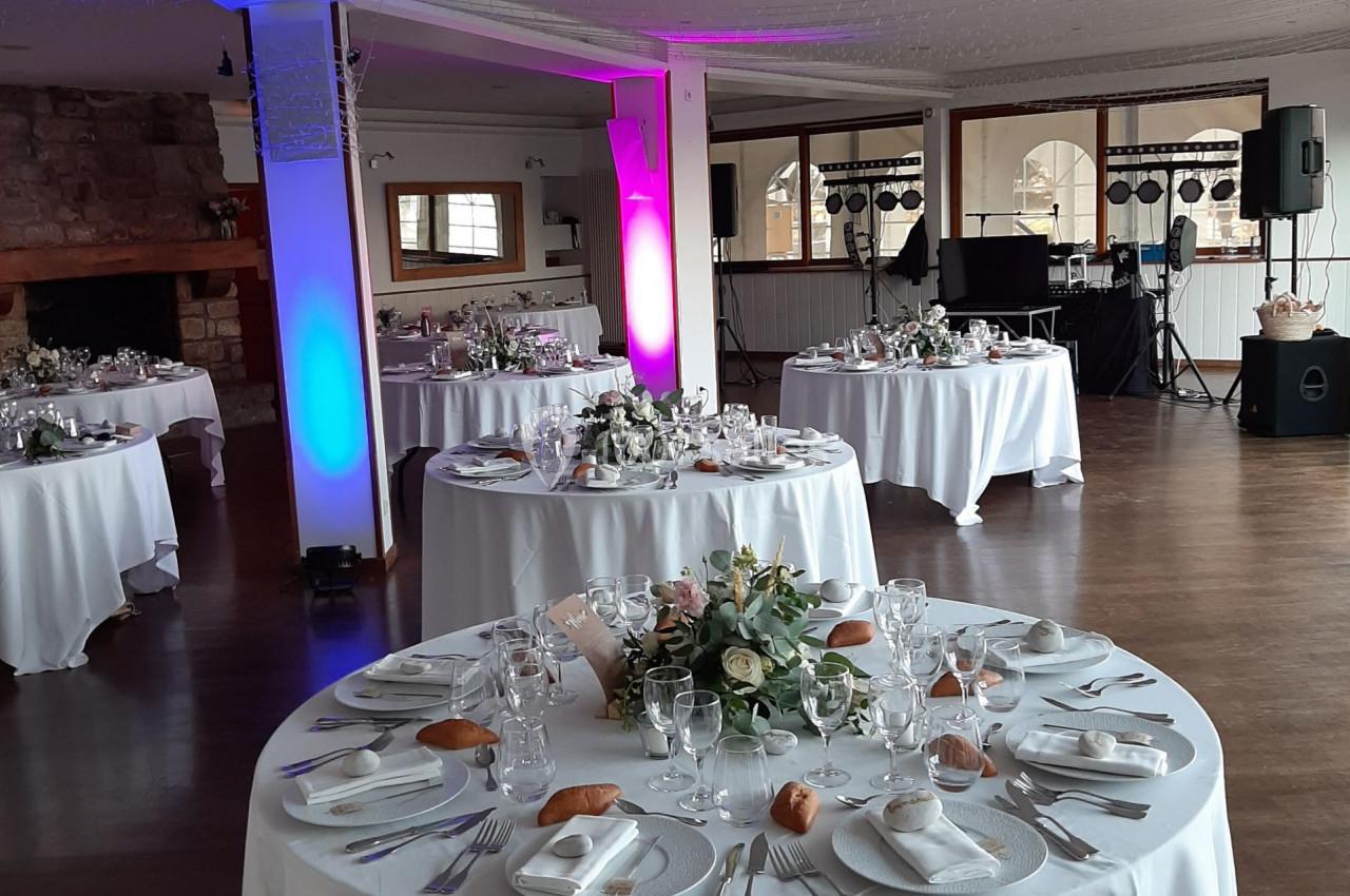 Salle de réception décorée avec des tables rondes dressées, éclairage coloré et équipement audio en arrière-plan.
