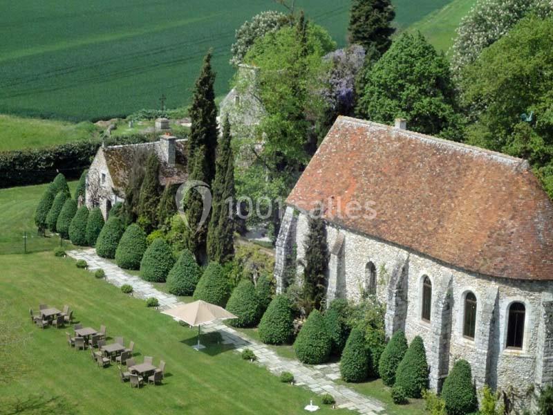 Vue aérienne d'un jardin aménagé avec des haies taillées, des tables, des parasols et des bâtiments en pierre au toit en…