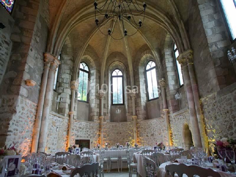 Salle voûtée en pierre avec de grandes fenêtres gothiques, décorée pour un événement avec tables dressées.