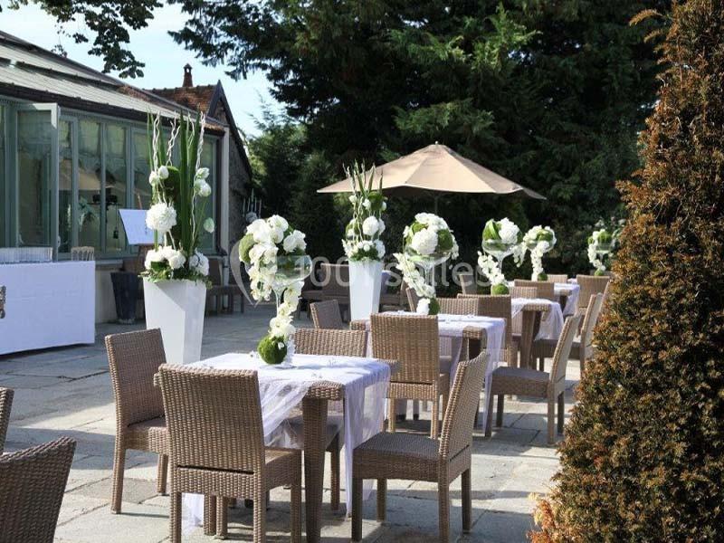 Terrasse aménagée avec des tables et chaises en rotin, décorée de grandes compositions florales blanches.