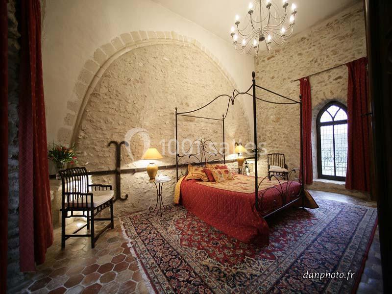 Chambre avec murs en pierre, lit à baldaquin, tapis coloré, chandelier et fenêtre en ogive.