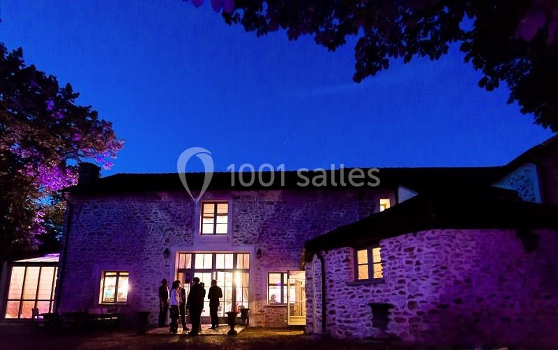 Façade d'une maison en pierre éclairée de nuit, avec des silhouettes de personnes discutant devant.