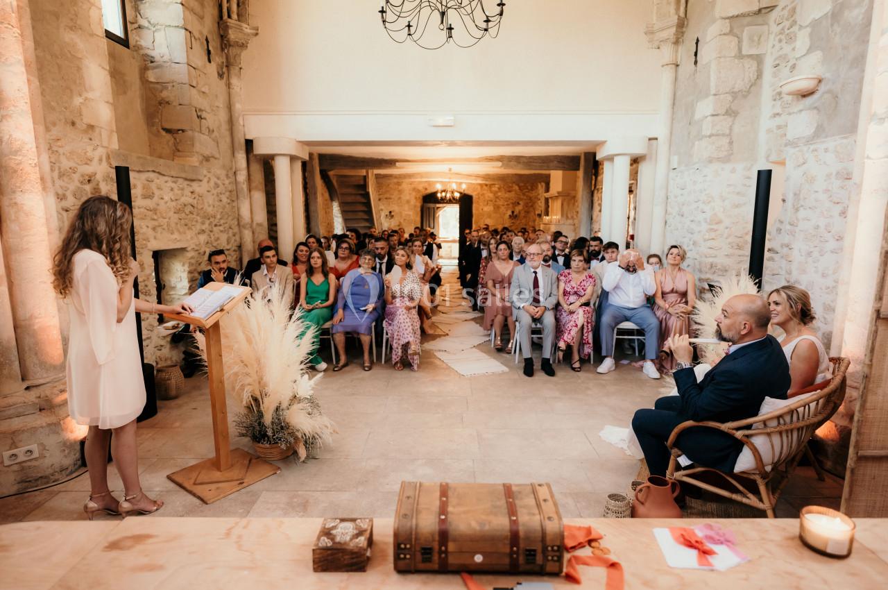 Une cérémonie dans une salle en pierre avec une officiant devant un public assis, décorée de fleurs et pampas.