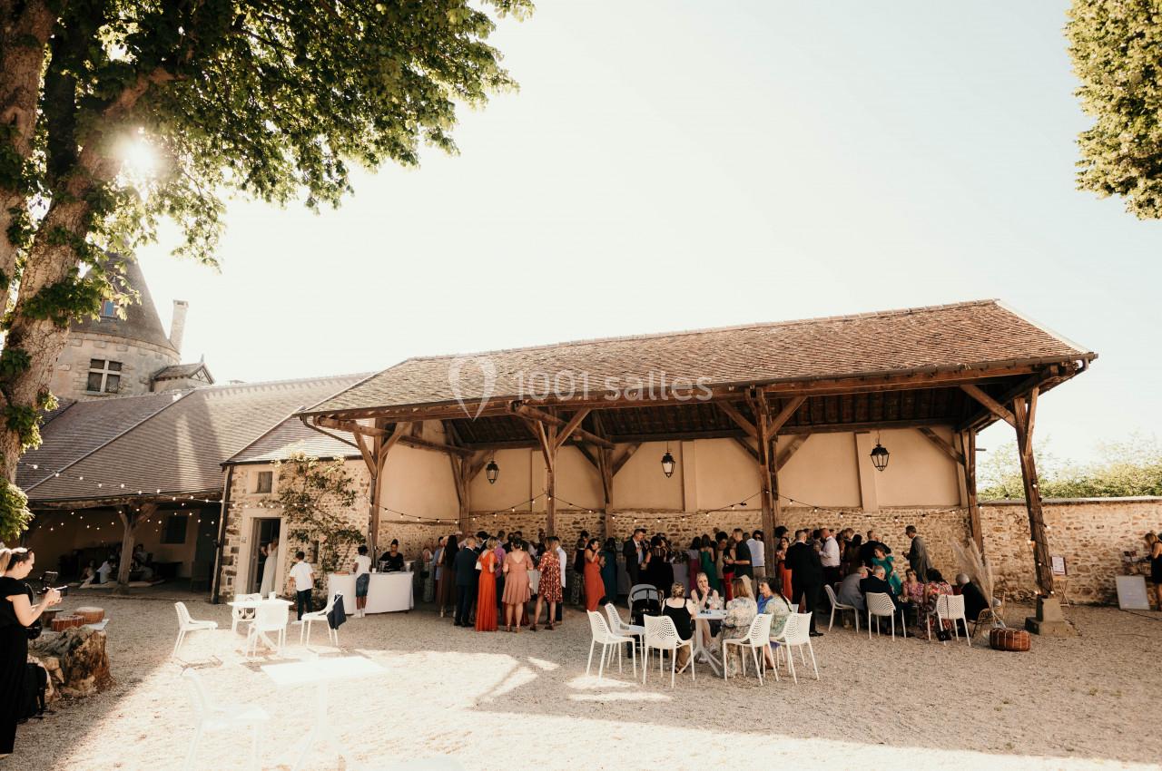 Groupe de personnes rassemblées sous une grande structure en bois dans une cour ensoleillée, avec des arbres et un bâtiment…