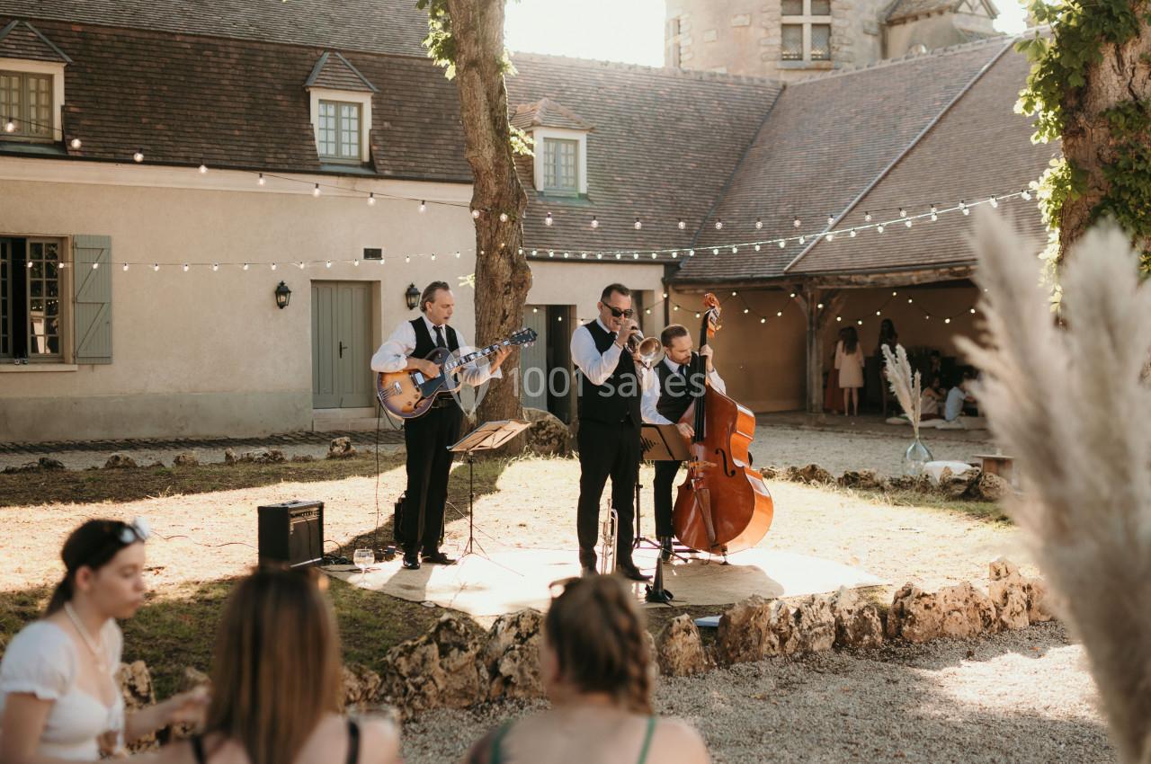 Un groupe de musiciens joue en plein air devant un bâtiment en pierre, avec des invités assis au premier plan.