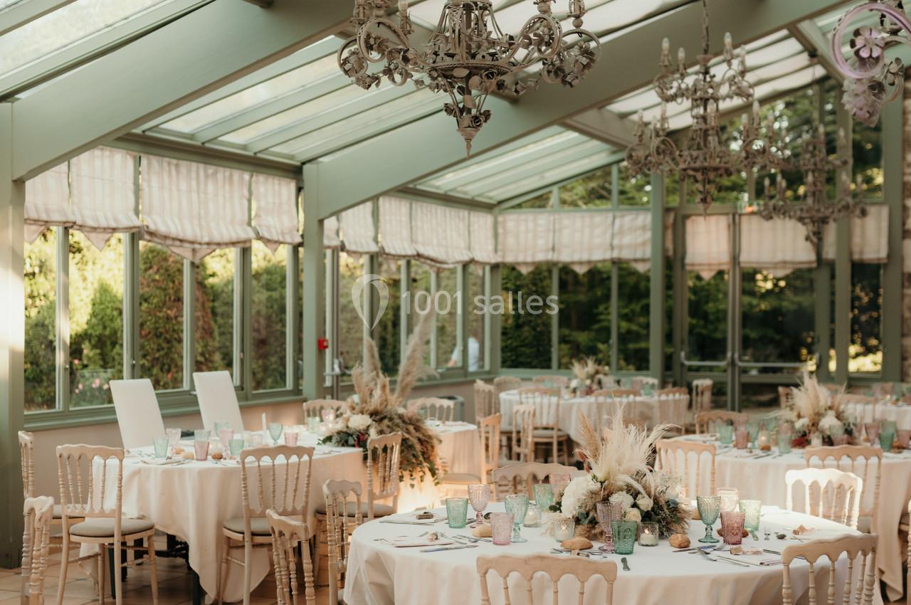 Salle lumineuse avec verrière, tables rondes dressées pour un événement, décorations florales et chandeliers suspendus.