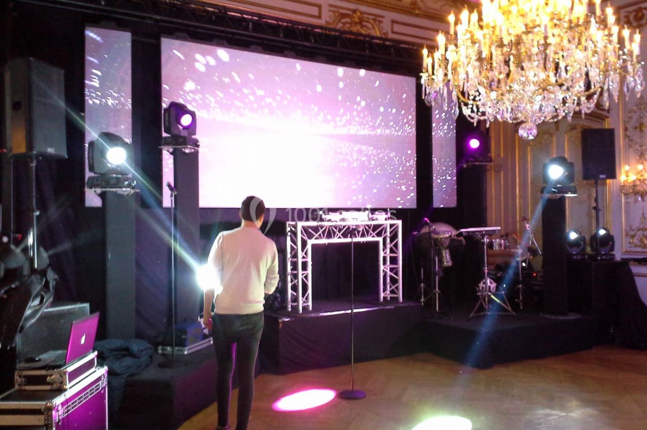 Homme debout dans une salle élégante avec écran géant, projecteurs colorés et lustre imposant.