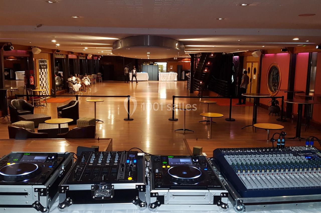 Salle spacieuse avec tables et chaises, vue depuis une régie équipée de platines DJ et d'une console de mixage.