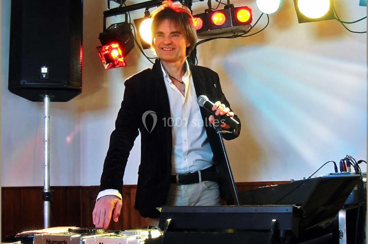 Un DJ souriant tenant un micro, debout derrière une table de mixage, éclairé par des projecteurs colorés.