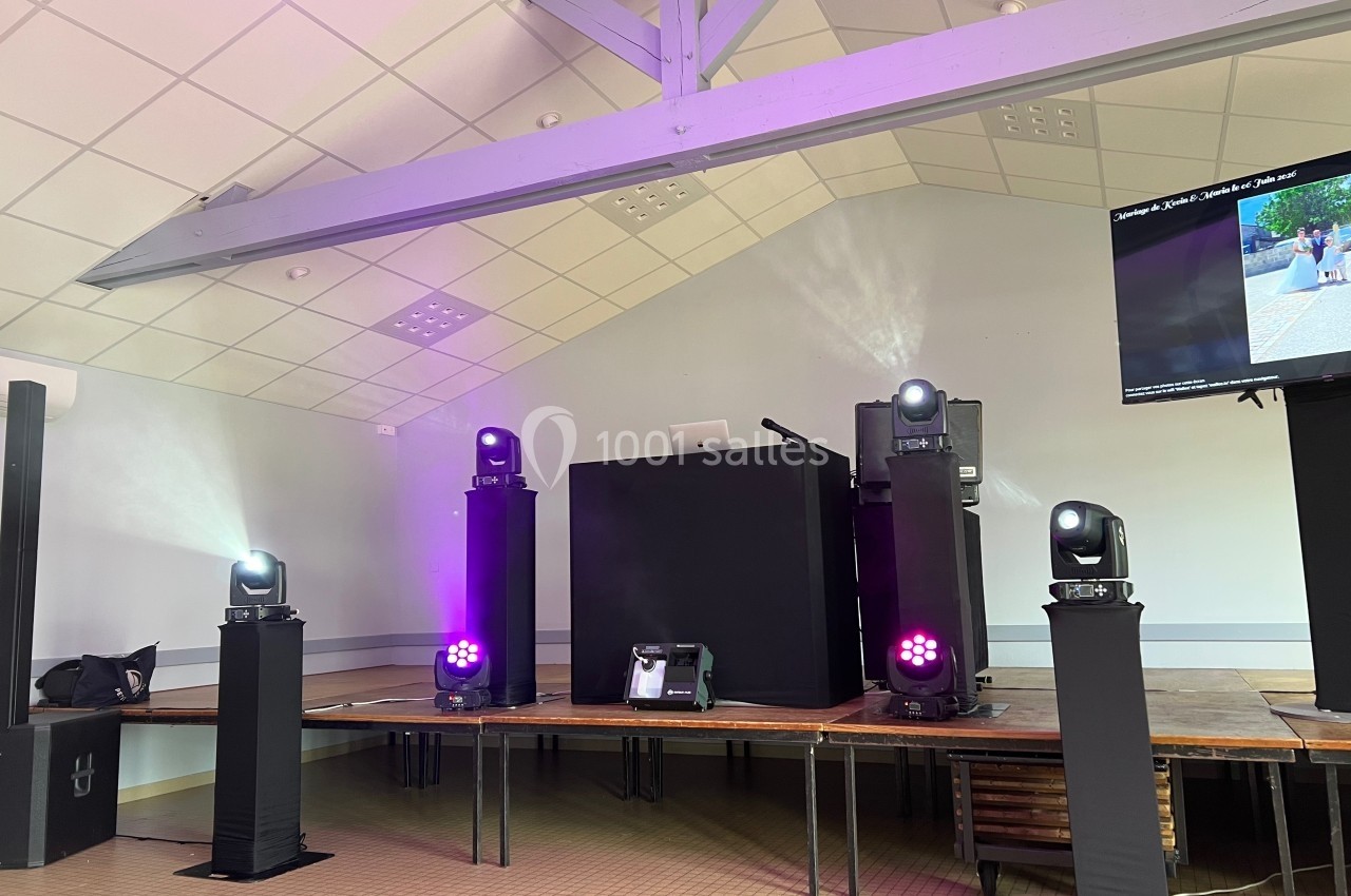 Scène équipée de projecteurs lumineux, d'une table de mixage et d'un écran dans une salle au plafond en pente.