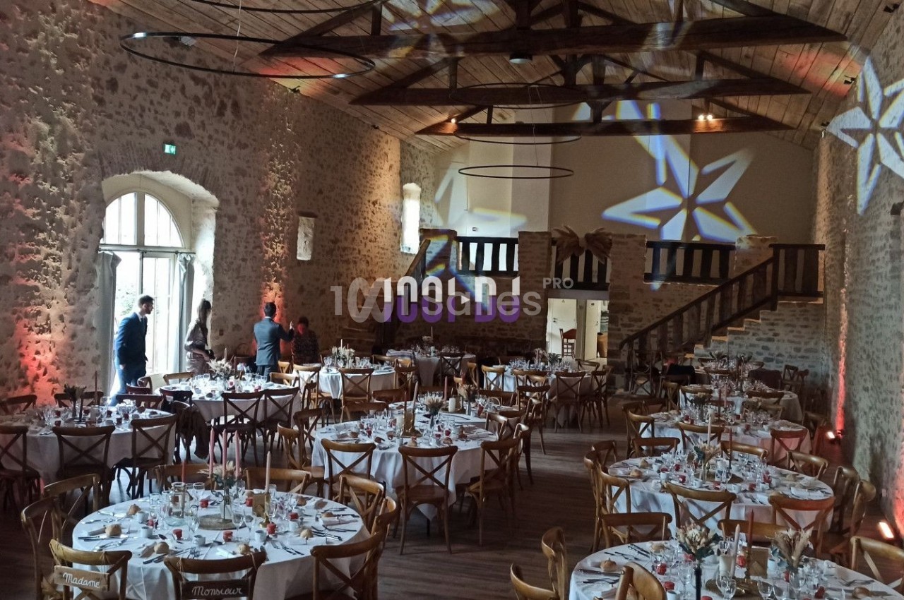 Salle de réception en pierre avec tables rondes dressées, éclairages décoratifs et mezzanine en bois.
