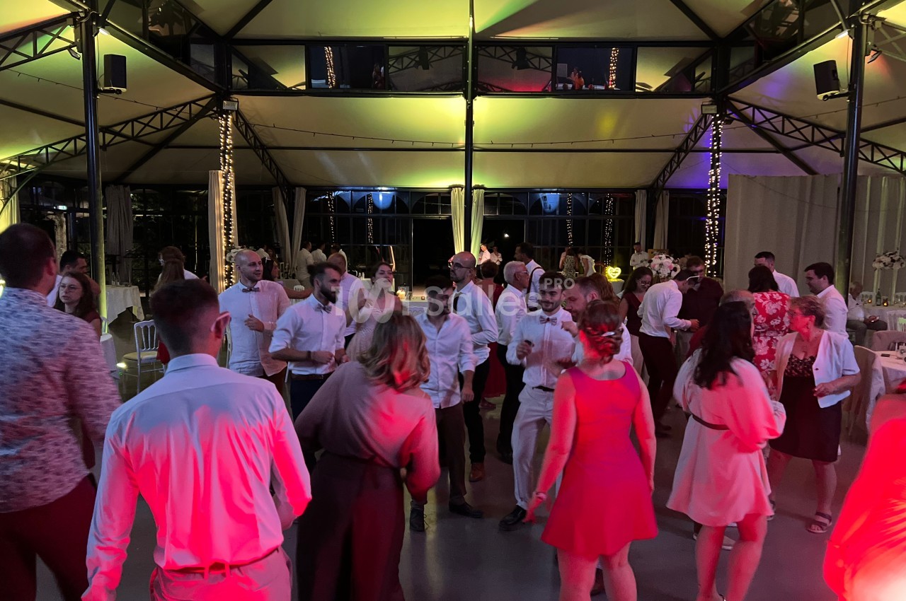 Des personnes dansent dans une salle éclairée de lumières colorées, entourées de tables décorées pour un événement.