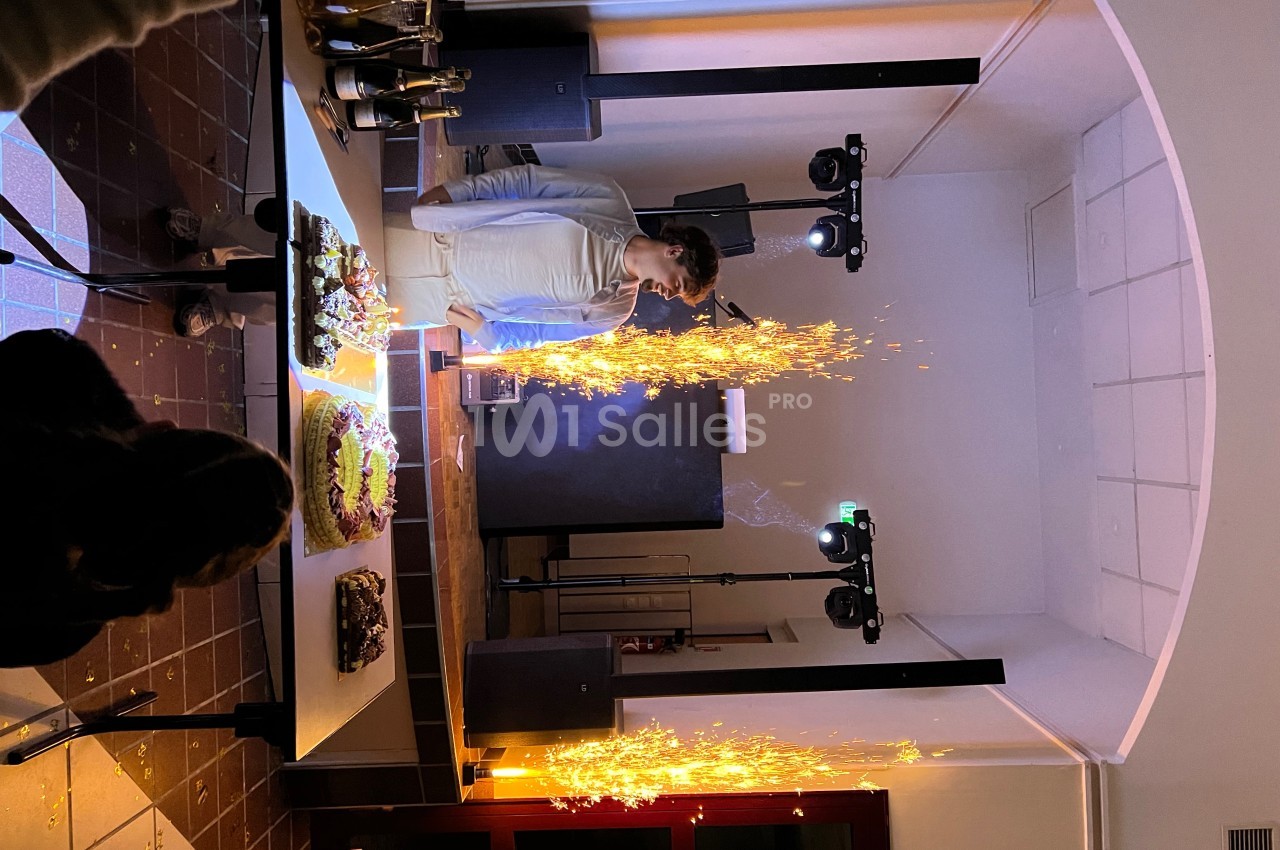 Un homme souffle des bougies sur un gâteau entouré de fontaines lumineuses dans une salle éclairée.