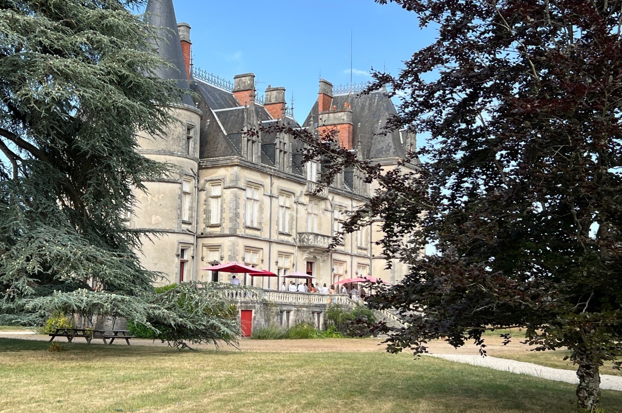 Château ancien avec tourelles, entouré d'arbres et d'une pelouse, sous un ciel bleu clair.