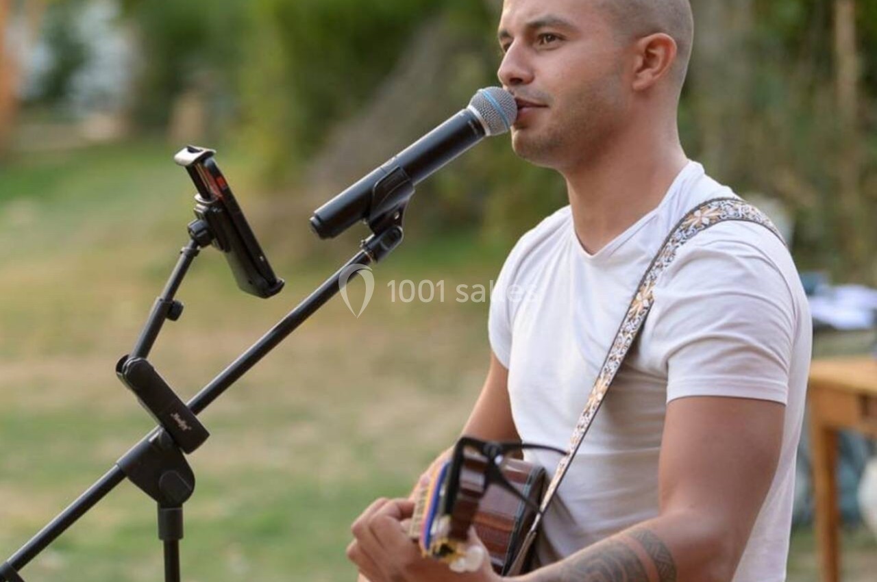 Un homme joue de la guitare et chante au micro en plein air, avec un support pour tablette à côté.