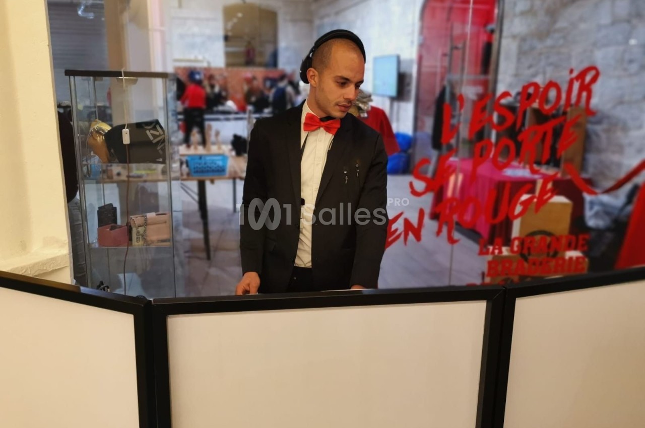 Un DJ en costume noir et nœud papillon rouge mixe devant un stand dans un espace intérieur.