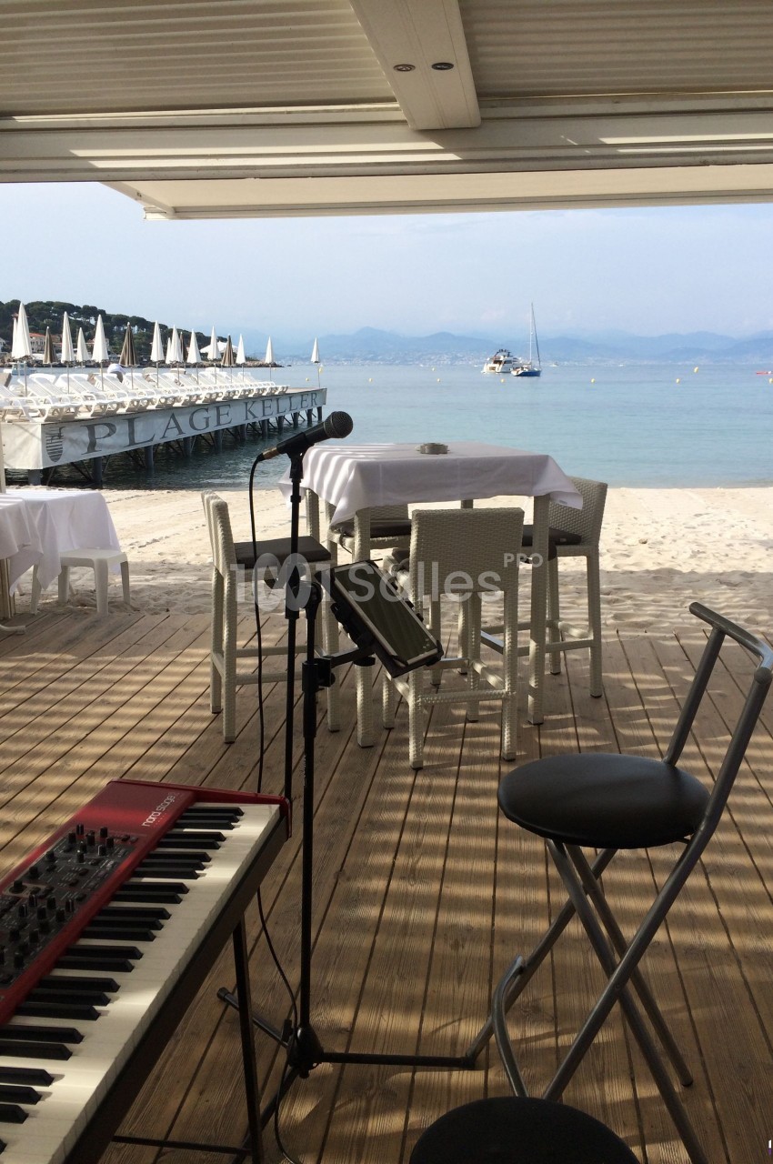 Vue d'une scène en bord de plage avec un clavier, un micro, des tables dressées et des bateaux au loin.
