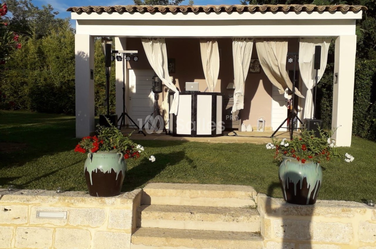 Pergola blanche avec rideaux ouverts, abritant une installation DJ, entourée de verdure et de pots de fleurs.