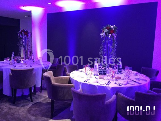 Salle de réception avec tables rondes dressées, décor floral et éclairage violet tamisé.