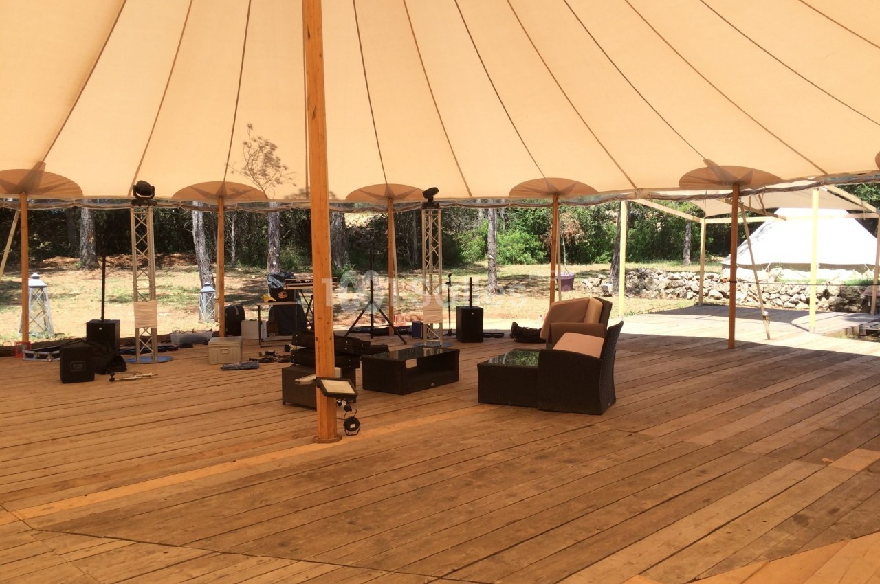 Intérieur d'une tente avec plancher en bois, mobilier d'extérieur et matériel technique, entourée de végétation.