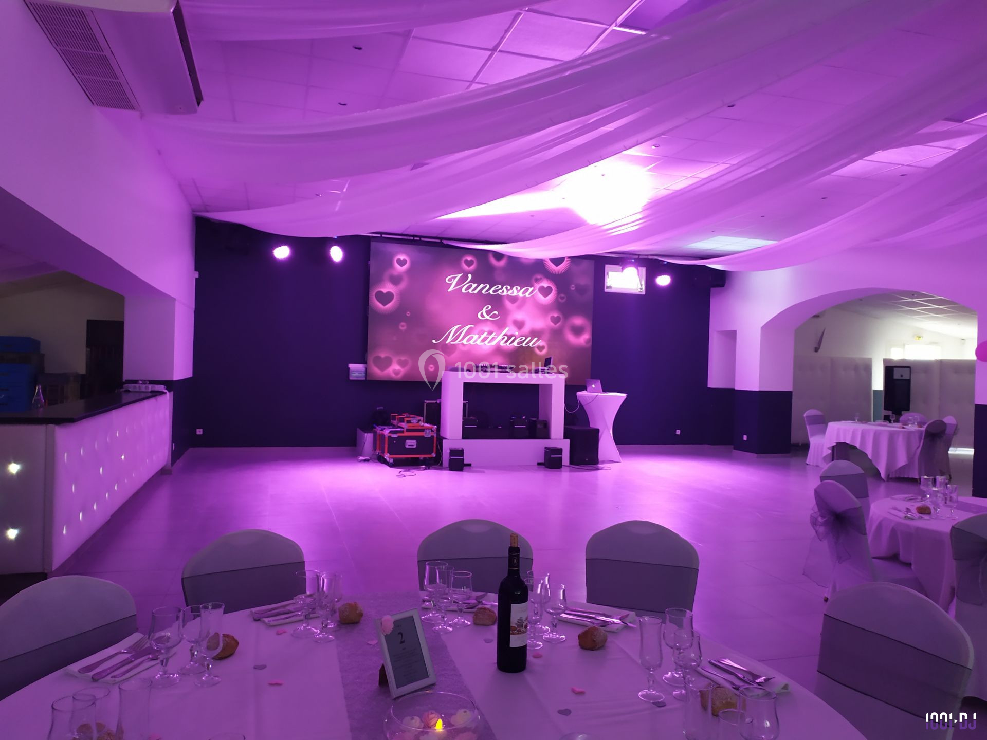 Salle de réception décorée avec des lumières violettes, tables dressées et écran affichant des prénoms.