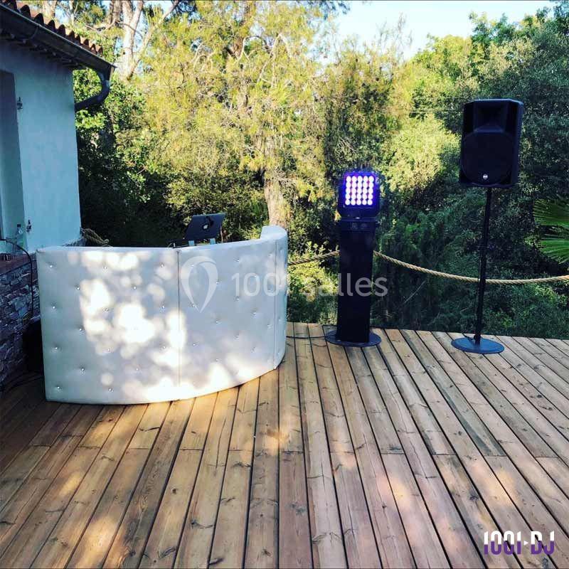 Console DJ blanche, enceinte et éclairage LED installés sur une terrasse en bois entourée de végétation.