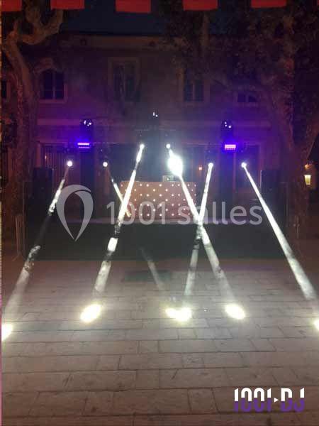 Scène extérieure éclairée par des projecteurs et faisceaux lumineux, avec une table de DJ en arrière-plan.